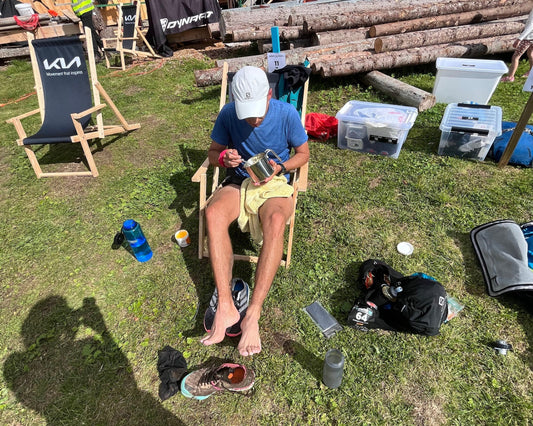 Ein Mann in sportlicher Kleidung sitzt auf einem Campingstuhl, isst aus einem Metallbecher und hat seine Laufschuhe ausgezogen. Neben ihm stehen Ausrüstungsgegenstände und Plastikboxen auf einer Wiese.