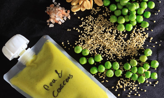 Eine wiederverwendbare Quetschflasche mit gelber Mischung, beschriftet mit 'Pea & Couscous', liegt auf einer schwarzen Unterlage. Daneben sind grüne Erbsen, Couscous, Erdnüsse und rosa Kristallsalz verstreut.