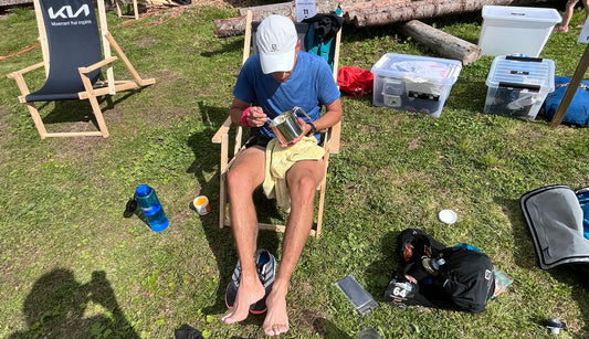 Ein Mann in sportlicher Kleidung sitzt auf einem Campingstuhl, isst aus einem Metallbecher und hat seine Laufschuhe ausgezogen. Neben ihm stehen Ausrüstungsgegenstände und Plastikboxen auf einer Wiese.