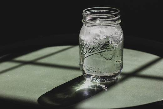 Ein durchsichtiges Einmachglas mit Wasser und Eiswürfeln, das auf einem Tisch mit Licht- und Schattenreflexionen steht.