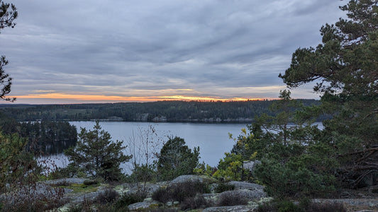 Eine malerische Landschaft mit einem ruhigen See, umgeben von Bäumen und Hügeln. Der Himmel zeigt einen sanften Sonnenuntergang mit orangefarbenem Schimmer unter einer Schicht grauer Wolken.