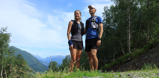 Eine Frau und ein Mann in sportlicher Laufkleidung stehen auf einem grasbewachsenen Pfad, umgeben von einer bewaldeten Landschaft und Bergen im Hintergrund unter blauem Himmel.