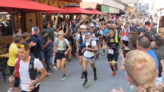 Eine Gruppe von Trailrunnern startet beim UTMB 2021, umgeben von jubelnden Zuschauern. Einige Läufer tragen Laufwesten und Startnummern, während Menschen in der Menge ihre Smartphones zum Fotografieren und Filmen hochhalten.