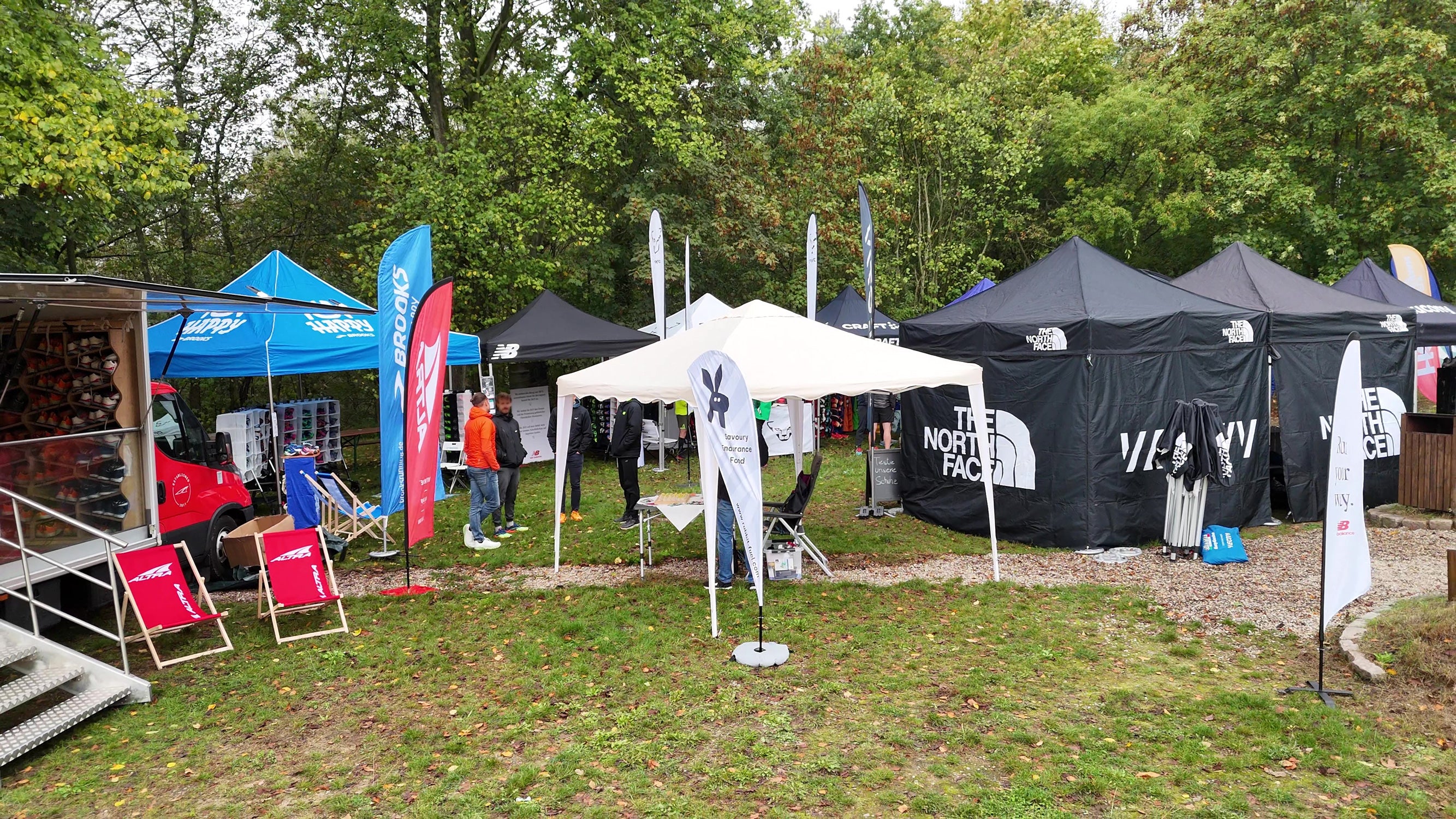 Eine Outdoor-Messe mit verschiedenen Markenständen, darunter RabbitFuel ‚The North Face, Brooks und Altra, die schwarze und bunte Pavillons aufgebaut haben. Vor einem roten Fahrzeug mit Verkaufsfläche stehen Liegestühle und Banner. Besucher schlendern zwischen den Ständen, die in einer grünen, bewaldeten Umgebung stehen.