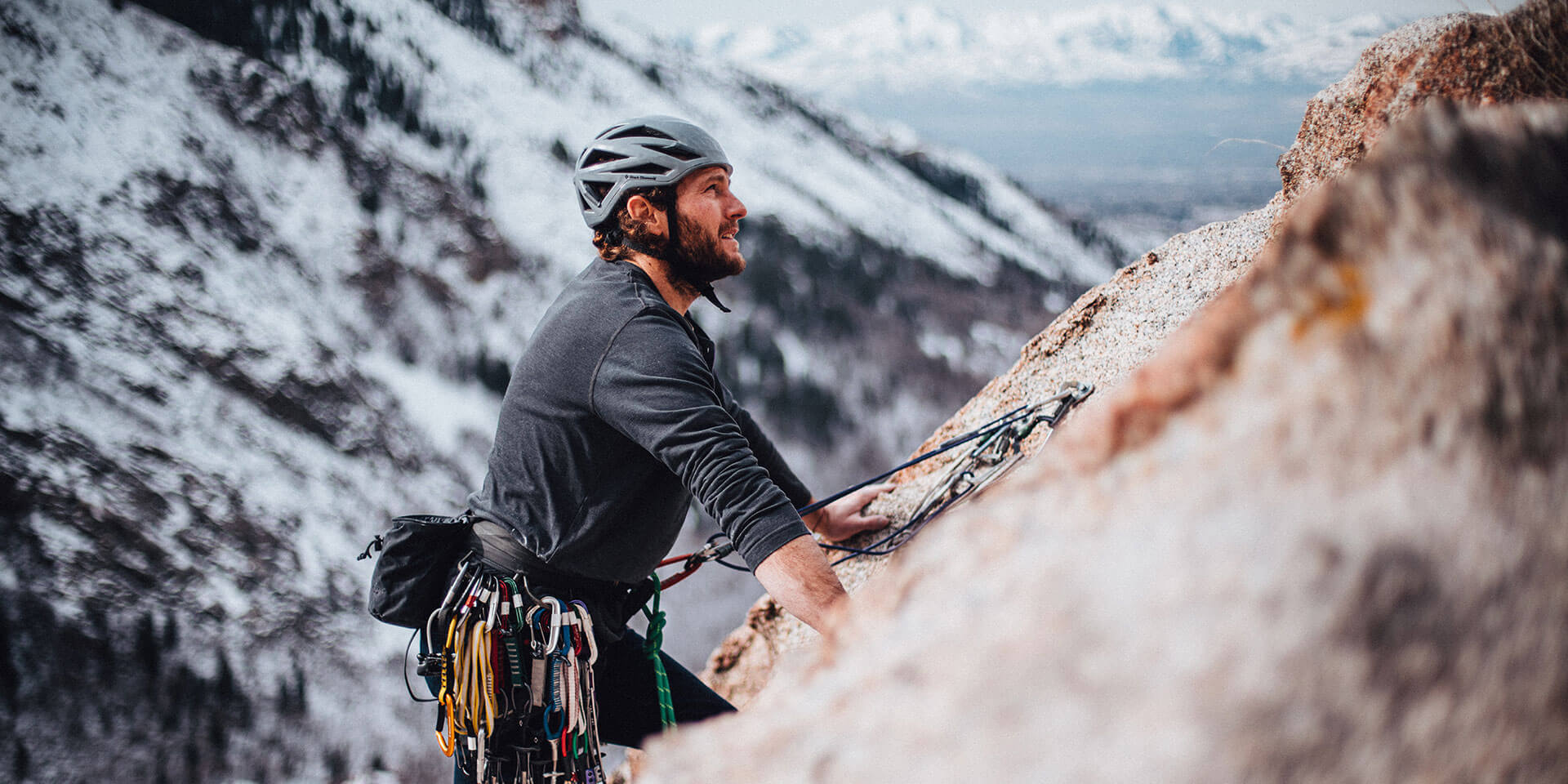 Ein Kletterer mit Helm und schwarzem Shirt erklimmt eine felsige Wand in einer verschneiten Berglandschaft. Er ist mit Seilen und Kletterausrüstung gesichert und blickt konzentriert nach oben. Im Hintergrund erstreckt sich eine winterliche Landschaft mit schneebedeckten Bergen.