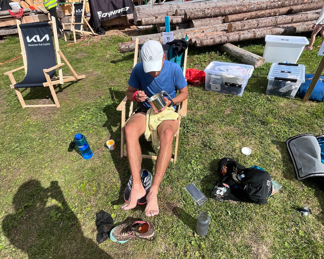 Ein Mann in sportlicher Kleidung sitzt auf einem Campingstuhl, isst aus einem Metallbecher und hat seine Laufschuhe ausgezogen. Neben ihm stehen Ausrüstungsgegenstände und Plastikboxen auf einer Wiese.