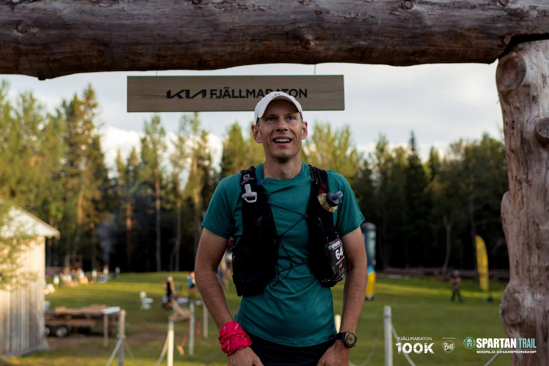 Ein Mann in sportlicher Kleidung steht unter einem Holzschild mit der Aufschrift 'KIA Fjällmaraton', umgeben von einer grünen, bewaldeten Landschaft und einer Wiese im Hintergrund.