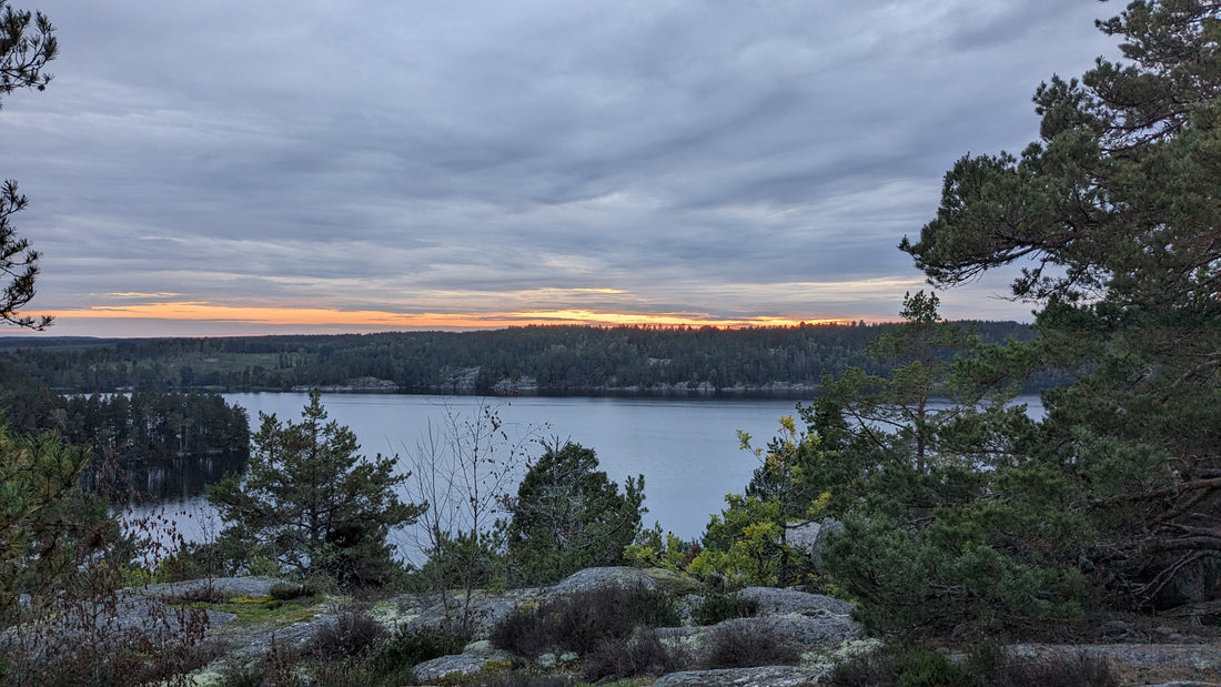 Eine malerische Landschaft mit einem ruhigen See, umgeben von Bäumen und Hügeln. Der Himmel zeigt einen sanften Sonnenuntergang mit orangefarbenem Schimmer unter einer Schicht grauer Wolken.