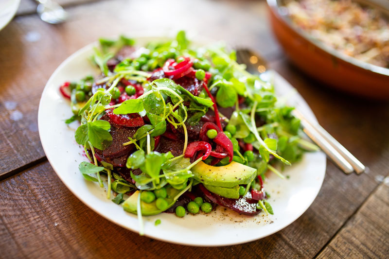 Ein Teller mit einem frischen Salat aus grünen Erbsen, Avocado, Rucola, roter Beete und Kräutern, angerichtet auf einem Holztisch.