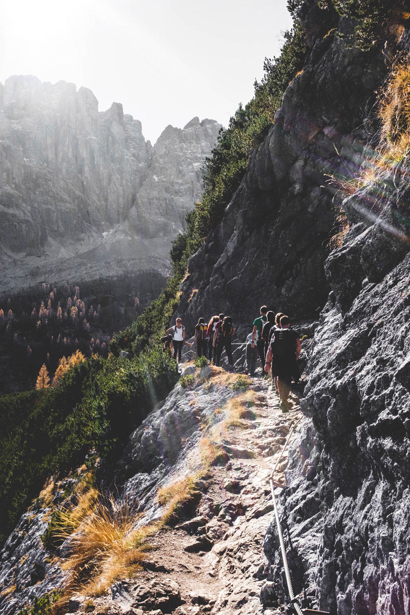 Eine Gruppe von Wanderern auf einem schmalen, felsigen Pfad, der an einem steilen Berghang entlangführt. Die Szene ist von Sonnenlicht durchflutet, und im Hintergrund sind beeindruckende Berggipfel sowie dichte Vegetation mit herbstlichen Farben zu sehen.