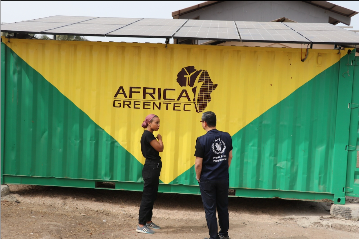 Zwei Personen stehen vor einem Container mit der Aufschrift ‚Africa GreenTec‘ und einer stilisierten Afrika-Karte. Der Container ist grün-gelb gestrichen und mit Solarpaneelen auf dem Dach ausgestattet. Die linke Person trägt schwarze Kleidung, die rechte Person eine Weste mit dem Logo des ‚World Food Programme‘. Die Szene befindet sich in einer sonnigen, trockenen Umgebung.