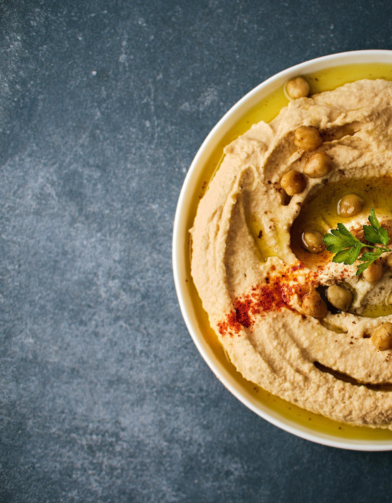 Nahaufnahme einer Schale mit cremigem Hummus, garniert mit Kichererbsen, Olivenöl, Paprikapulver und frischer Petersilie, auf einem dunklen Hintergrund.