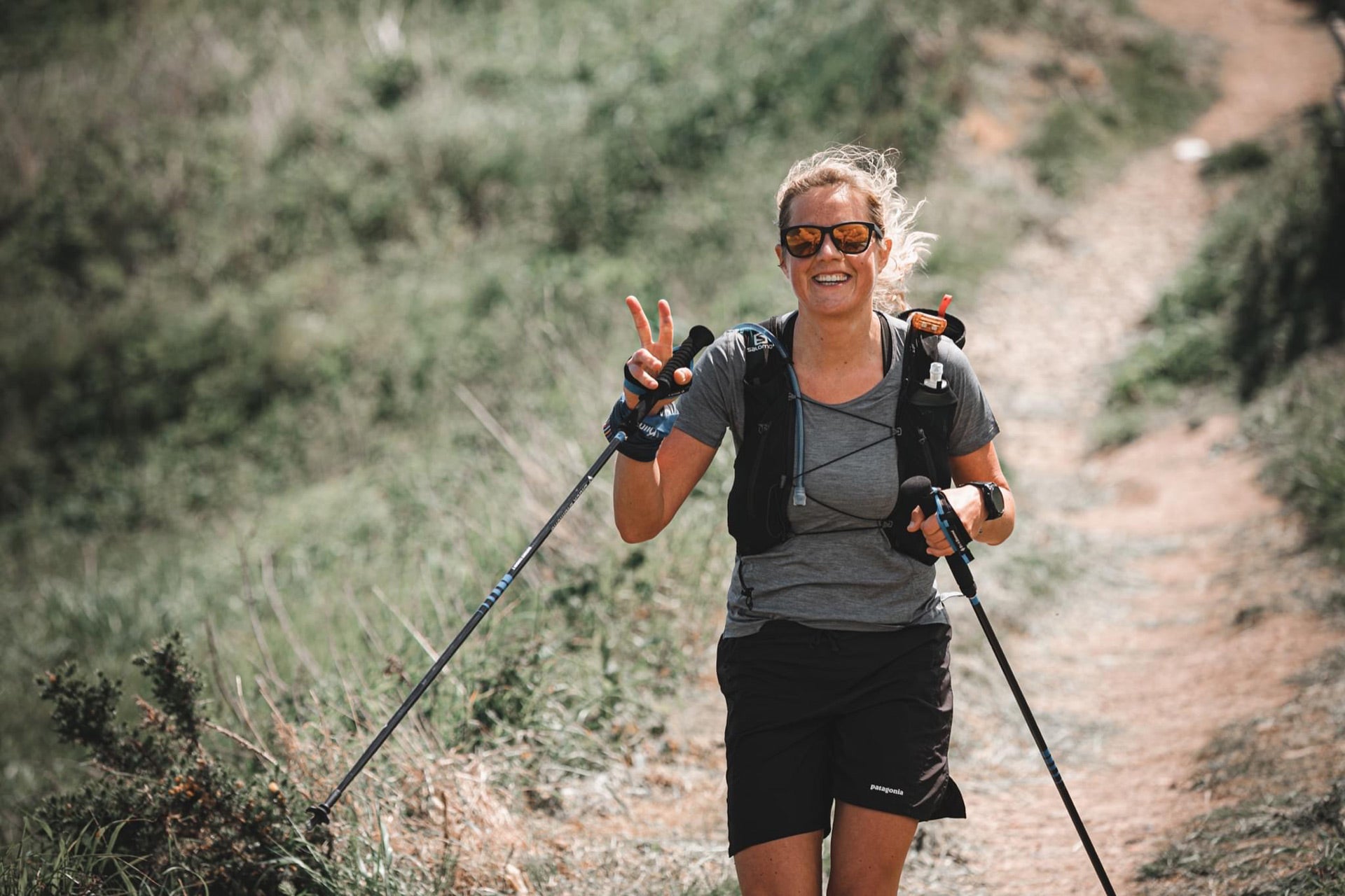 Eine Frau in sportlicher Kleidung wandert fröhlich mit Trekkingstöcken auf einem schmalen, von Gras und Büschen gesäumten Pfad. Sie trägt eine Sonnenbrille, eine schwarze Weste mit Trinkflaschen und hebt die Hand, um ein Friedenszeichen zu zeigen, während sie lächelt.