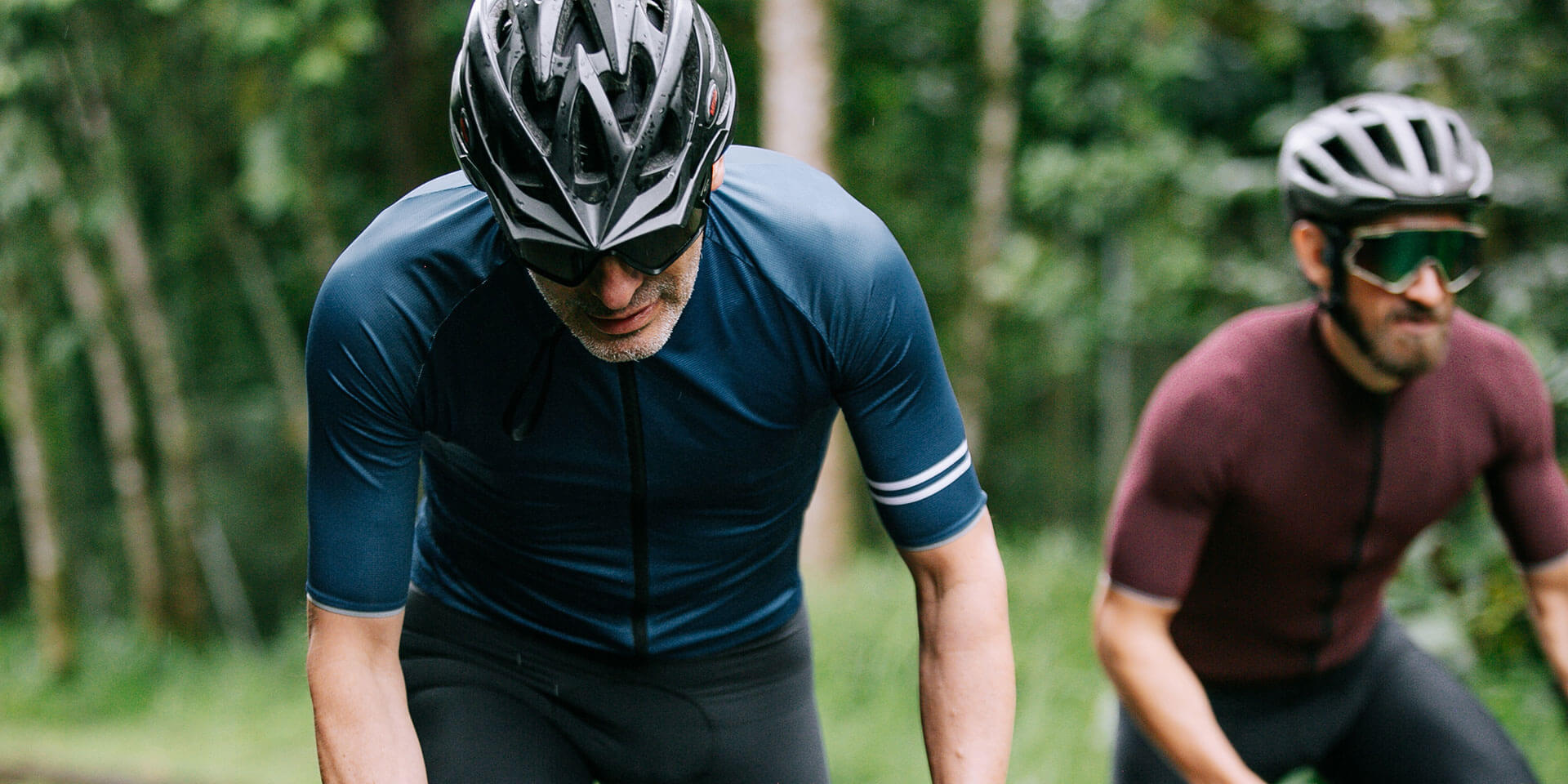 Zwei Radfahrer in enganliegenden Trikots und Helmen fahren konzentriert durch eine grüne, bewaldete Umgebung. Der vordere Fahrer trägt ein dunkelblaues Trikot mit weißen Streifen am Ärmel, der hintere Fahrer ein weinrotes Trikot. Beide tragen Sonnenbrillen und wirken auf den Sport fokussiert.